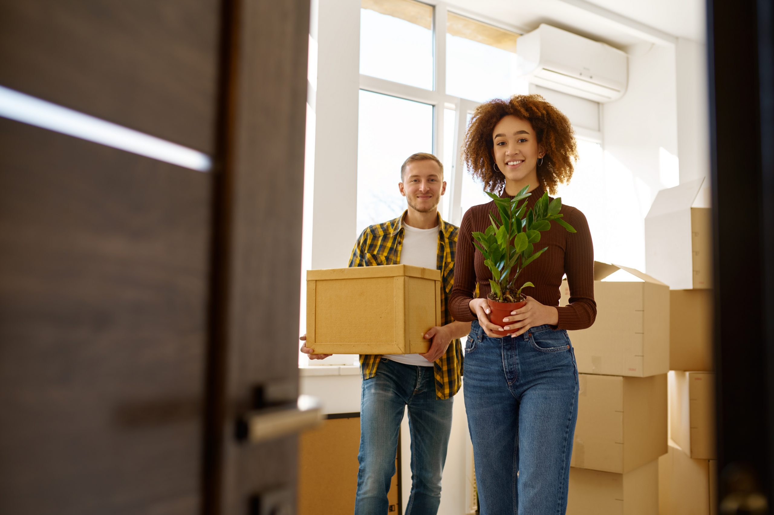 Happy young couple on moving day carrying cardboard boxes with belongings and flowers to illustrate Apartment Movers Near Me Portland, Vancouver